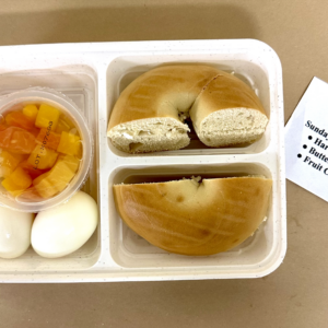 A Cook For Vets breakfast meal packed for delivery: hard-boiled eggs, a buttered bagel, and a fruit cup in a divided container